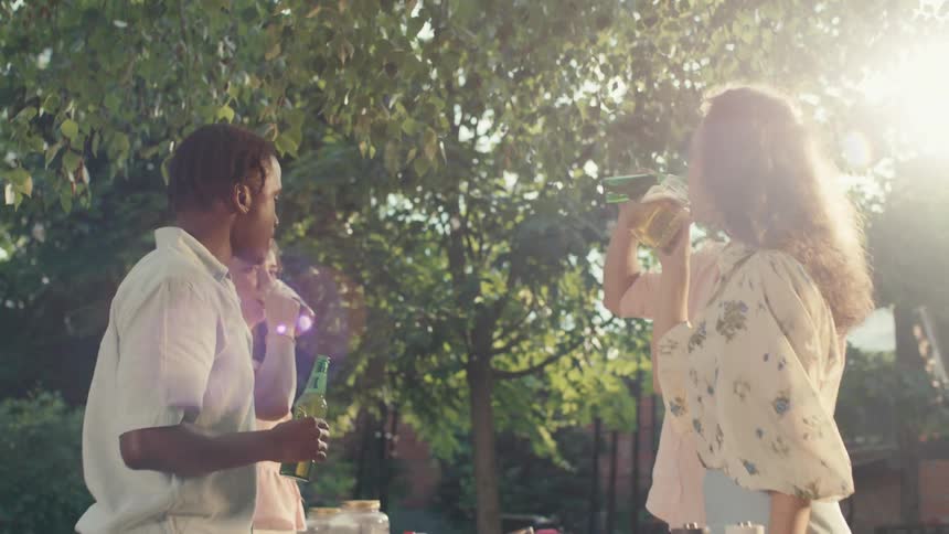 A diverse group of young friends toast with beers at a sunny backyard party, laughing and enjoying the summer day
