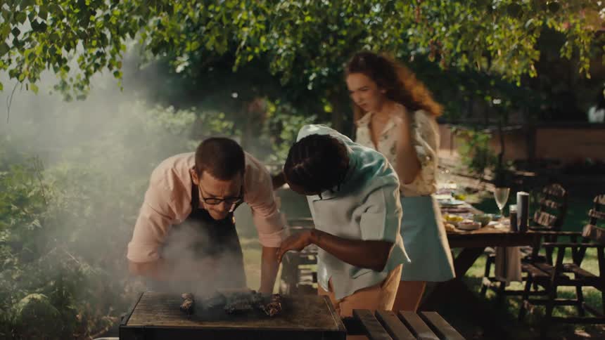 Two men playfully fan away excessive smoke from a grill during a summer backyard barbecue with friends