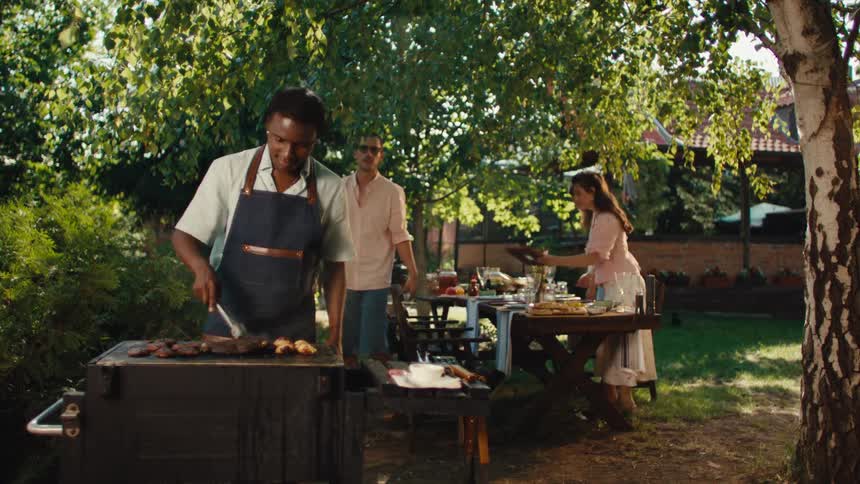 A diverse group of friends gathers for a summer barbecue in a sunny backyard, with one man grilling meat while others prepare the dining table