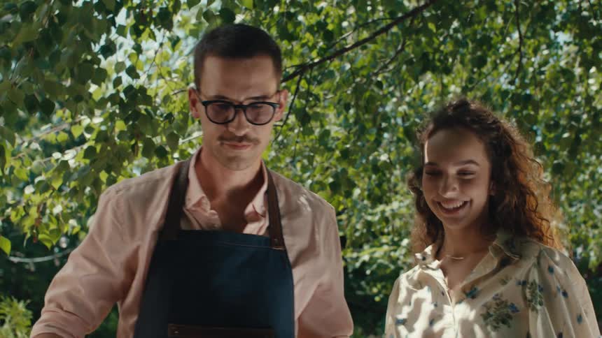 A low-angle slow-motion shot reveals a young couple smiling while barbecuing and drinking beer in a sunny backyard