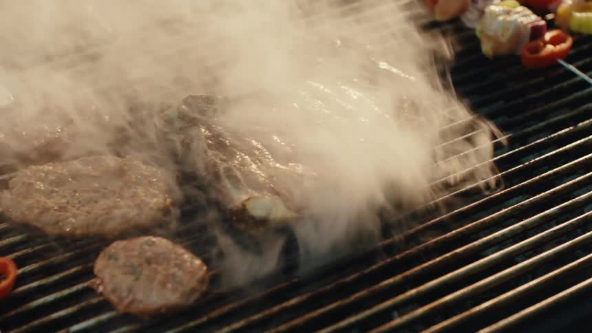 Close up slow motion shot of a juicy T-bone steak and burger patties being flipped on a smoking hot barbecue grill next to vegetable skewers