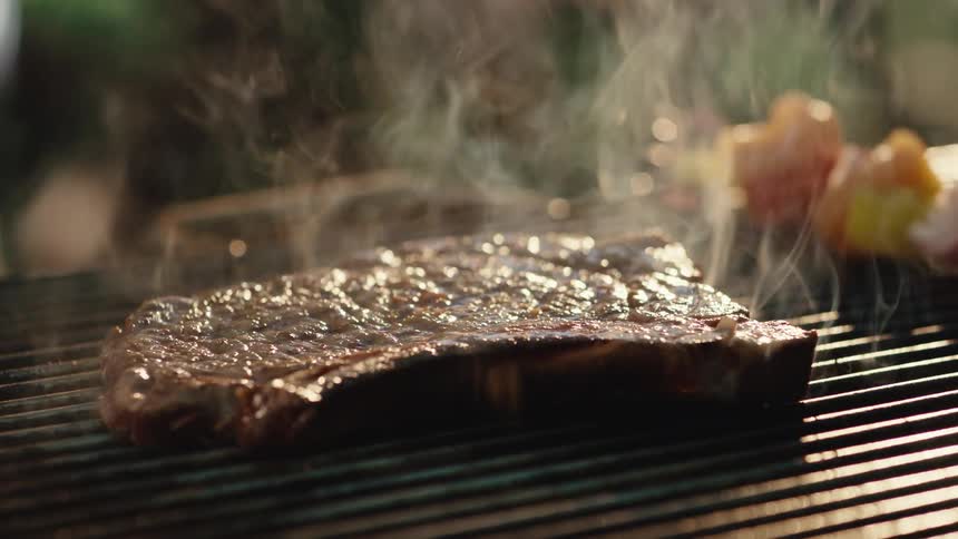 A juicy T-bone steak is placed on a hot smoking grill and flipped with tongs, causing flames to erupt in slow motion