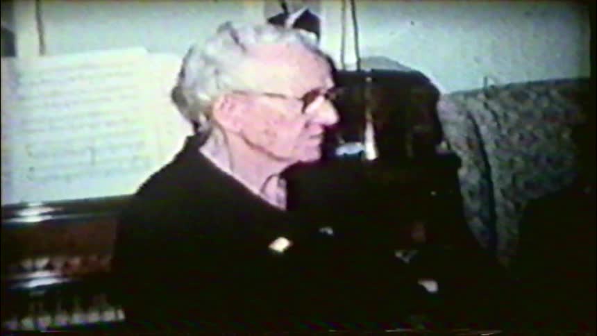 A camera-shy elderly man with glasses covers his face while sitting next to an organ in his home
