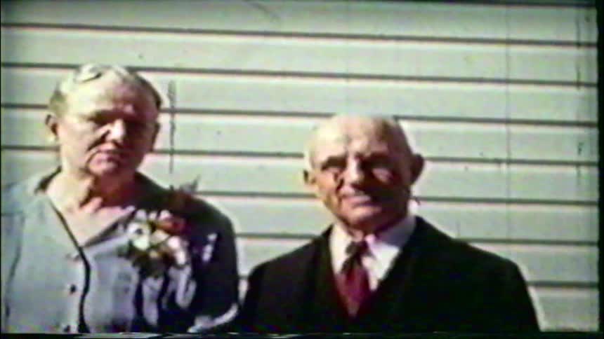 An old, grainy home movie shows an elderly couple posing for a portrait in front of a house