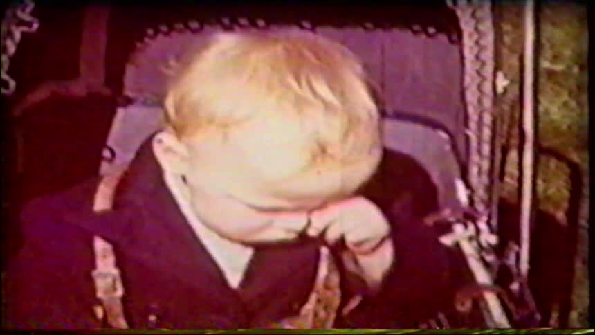 A vintage home movie captures a close-up of a blonde-haired toddler sitting in a stroller who begins to cry and rub their eyes