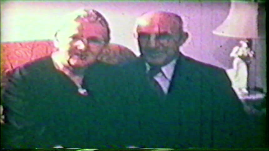 A vintage home movie captures a loving elderly couple smiling and talking while sitting on a sofa in their living room