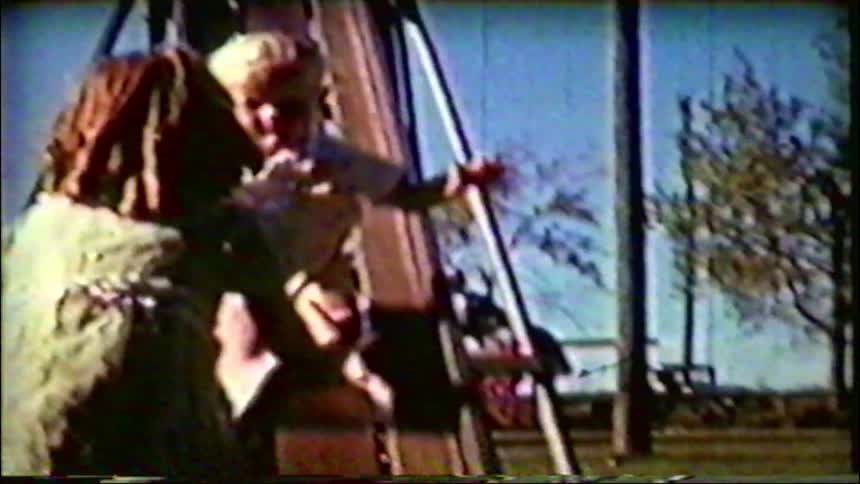 A toddler excitedly slides down a playground slide into the waiting arms of a family member in this charming vintage home movie