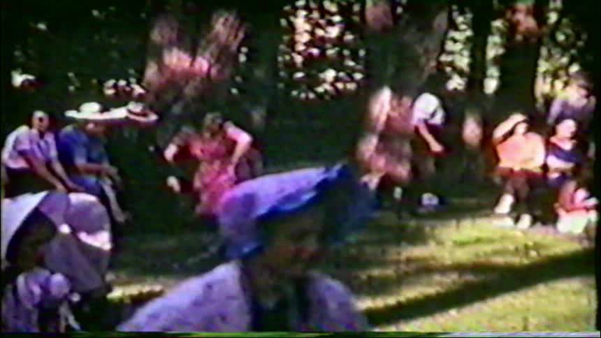 Grainy archival footage shows a group of women and children in traditional clothing playing games at a community picnic in a park