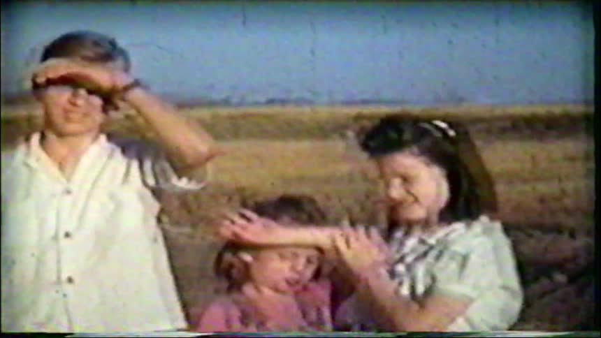 Vintage 8mm footage shows three siblings shielding their eyes from the sun in a rural field on a bright day