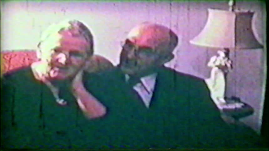 A nostalgic vintage home movie captures a sweet elderly couple sitting closely together on a sofa in their living room