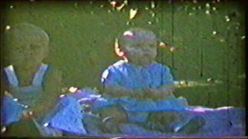 A grainy vintage home movie captures two babies sitting on a blanket on the grass on a sunny day