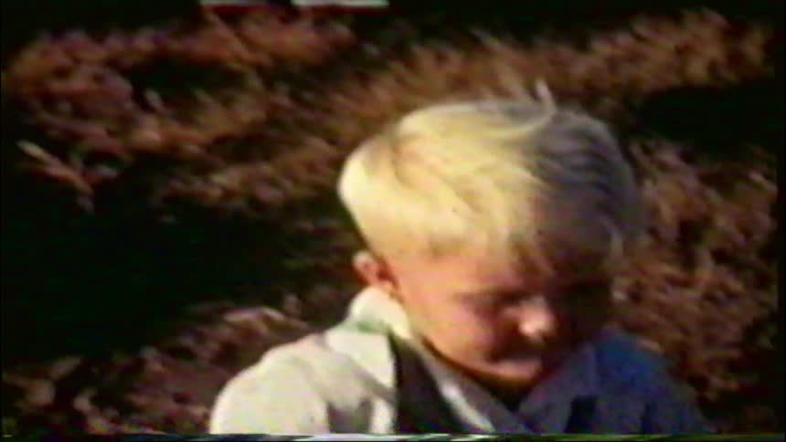 Vintage home movie captures a young blond-haired boy riding his bicycle outside in a yard