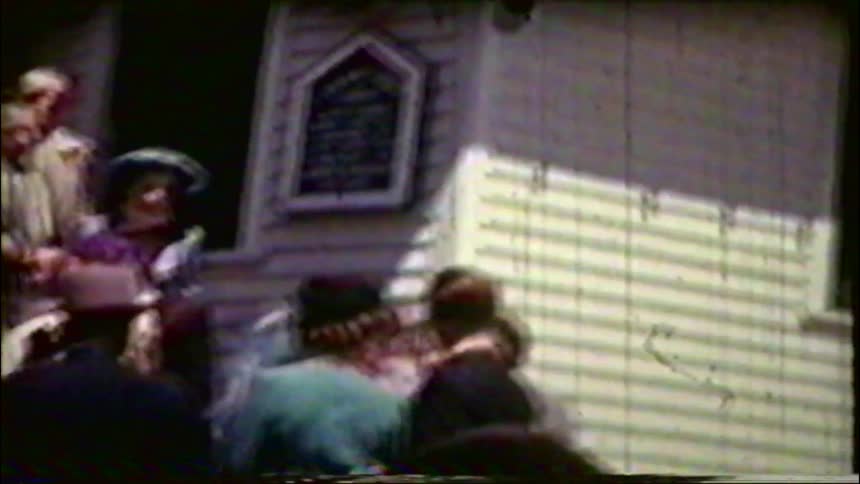 Archival footage shows a large crowd of well-dressed people socializing outside a building in the 1940s