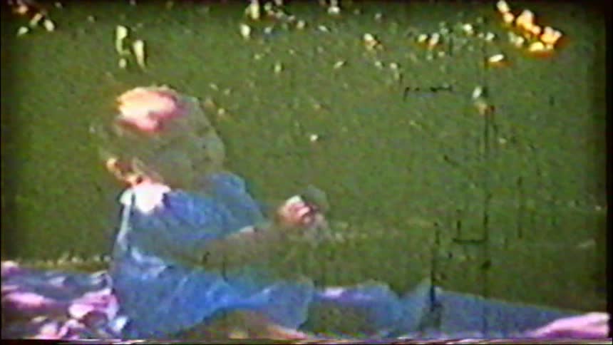 A grainy vintage home movie captures a sweet baby sitting on a blanket in a sunny, grassy backyard