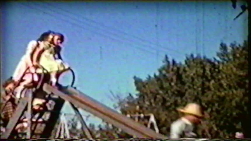 Nostalgic vintage footage shows a young boy in a straw hat sliding down a slide while two girls watch from the top