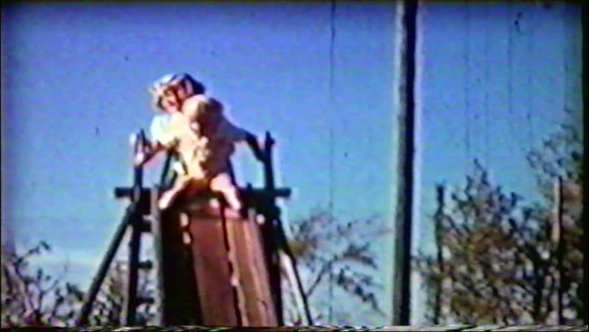 Vintage footage captures two young children hesitating at the top of a playground slide on a sunny day
