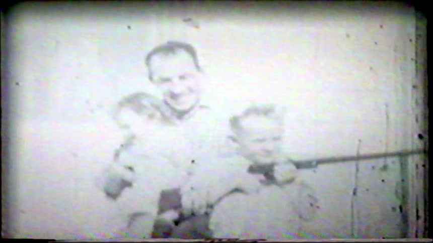 Vintage home movie footage shows a father kissing his toddler daughter and posing with his young son who is holding a toy rifle