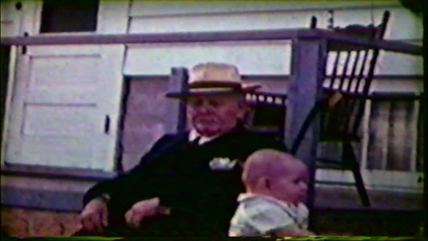 A grandfather in a suit and hat sits on a porch with his grandchild in this vintage home movie