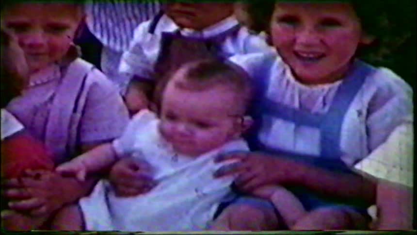 A group of adorable children and a baby smile and laugh together in a nostalgic vintage home movie