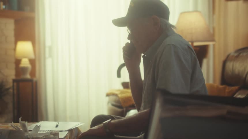 A senior man wearing a cap has a thoughtful conversation on the phone in his warm, sunlit living room
