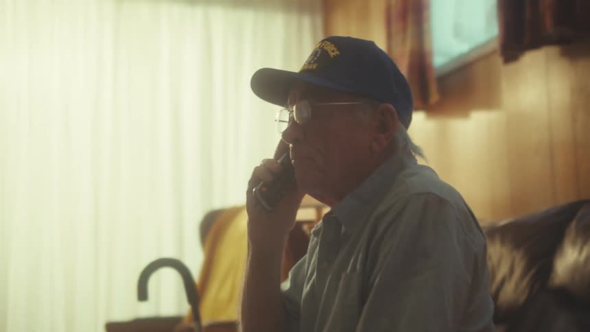 A senior Air Force veteran sits on his couch in a warmly lit living room while talking on a cordless phone