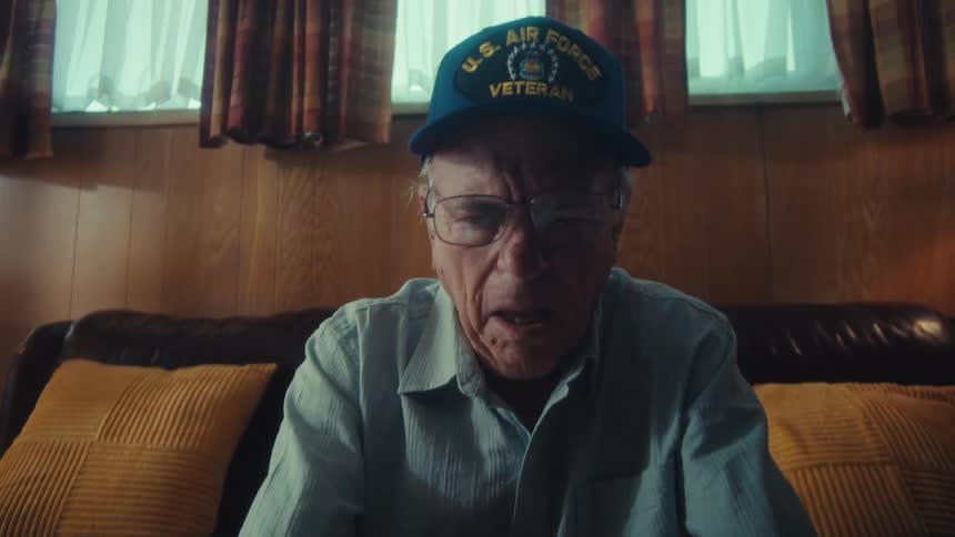 An elderly U.S. Air Force veteran sits on his couch looking stressed and overwhelmed while sorting through a large pile of bills and receipts in his living room