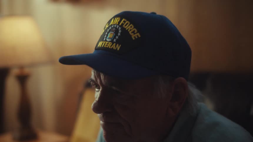 A solemn, elderly Air Force veteran removes his glasses while sitting in quiet reflection in a dimly lit room