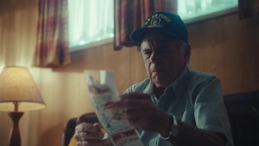 An elderly Air Force veteran with a thoughtful expression reads a map in his dimly lit home, looking pensive and concerned
