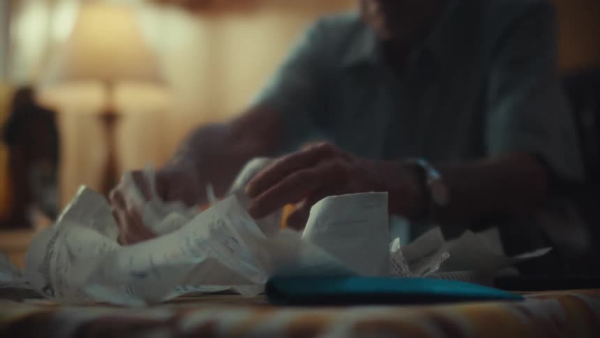 A stressed elderly Air Force veteran sits at his table overwhelmed as he sorts through a large pile of bills and receipts
