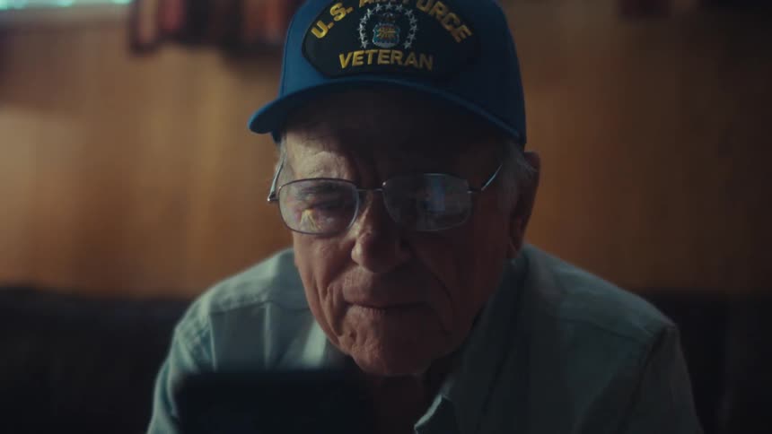 A slow push-in shot reveals an elderly U.S. Air Force veteran sitting on a couch, looking intently at his smartphone in a dimly lit living room