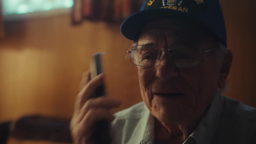 A somber elderly veteran wearing an Air Force hat hangs up the phone with a sad expression while sitting alone in his living room