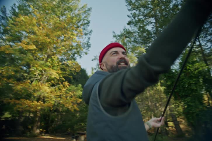 A smiling man in a red beanie sets up a tent in a forest clearing during golden hour