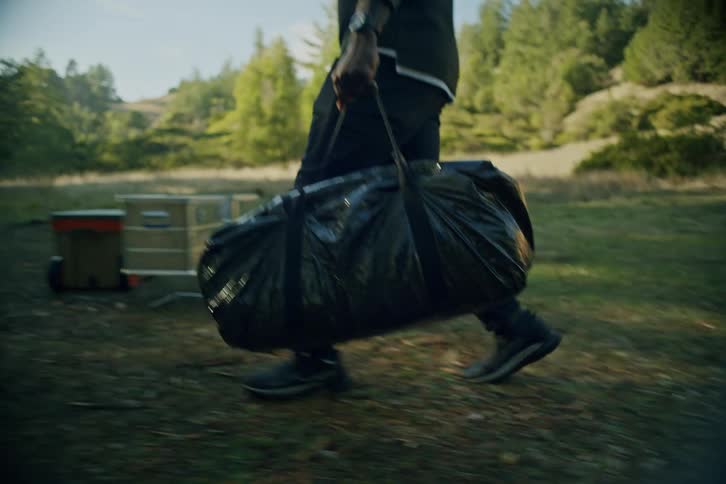 A person carries a large black duffel bag through a grassy field to set up camp