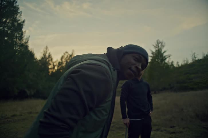 Two diverse friends stand in a serene forest clearing at dusk, with one man turning to look directly at the camera