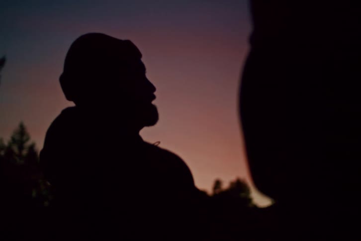 Silhouette of a man with a beard and beanie talking to a friend against a beautiful twilight sky