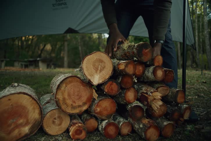 A camper carries a bundle of chopped logs and adds them to a firewood pile in slow motion at a campsite