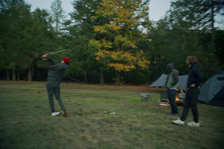 A group of male friends enjoy a casual game of golf while camping in a beautiful forest clearing at dusk