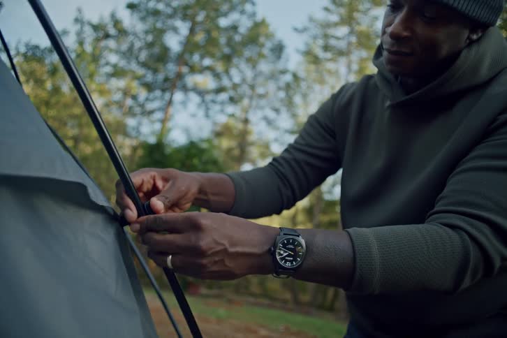 A man in a beanie and hoodie carefully assembles a tent, clipping the fabric to the poles in a wooded campsite