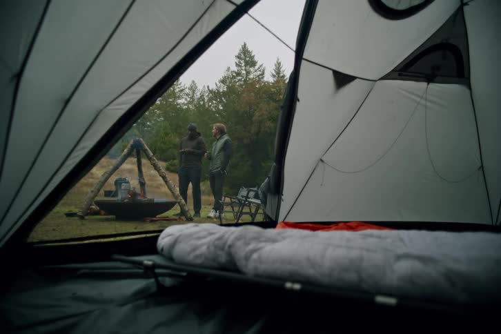 Two friends relax by a campfire on a camping trip, viewed from inside a cozy tent on a cloudy day
