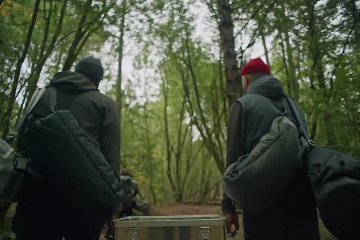 Rear view of two men carrying a heavy metal case and camping gear as they walk through a dense forest