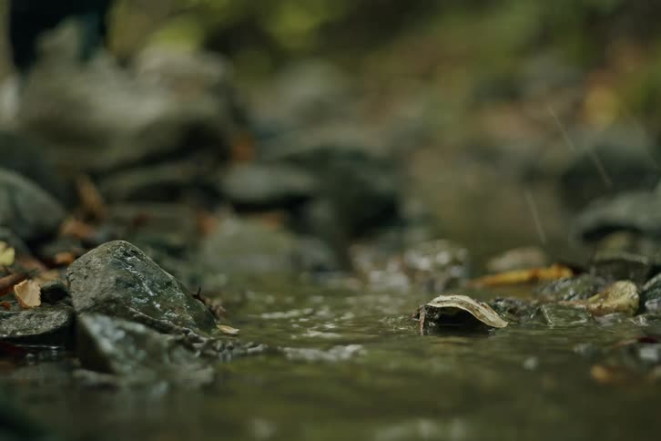 A slow-motion close-up of various hiking shoes and boots stepping through a rocky stream, splashing water with each step