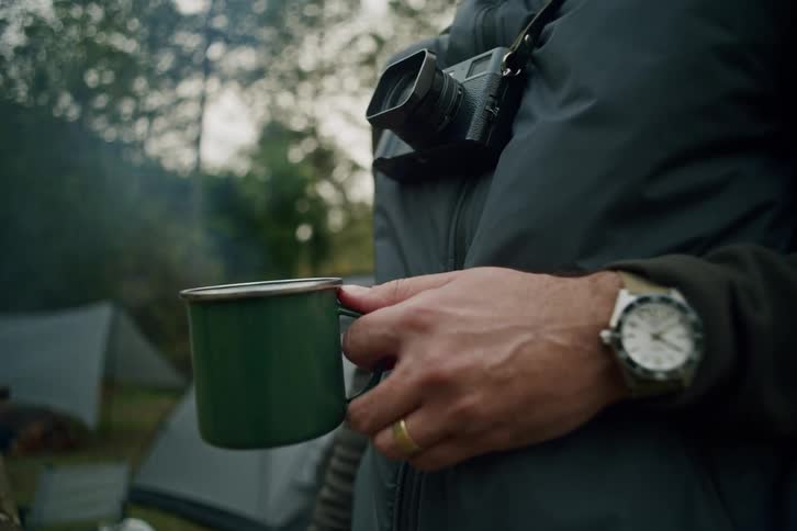 Close-up on a man with a camera holding a steaming hot drink in a green mug at his campsite