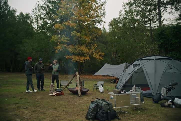 Three male friends gather around a smoking campfire, sharing stories and enjoying a relaxing morning at their wooded campsite