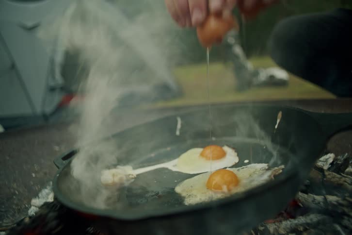 A person cracks eggs into a hot, steaming cast iron skillet over a campfire for a rustic breakfast in slow motion