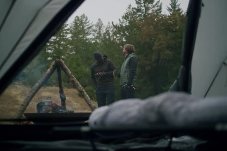 A view from inside a tent shows two male friends chatting and drinking coffee by a warm campfire at their campsite