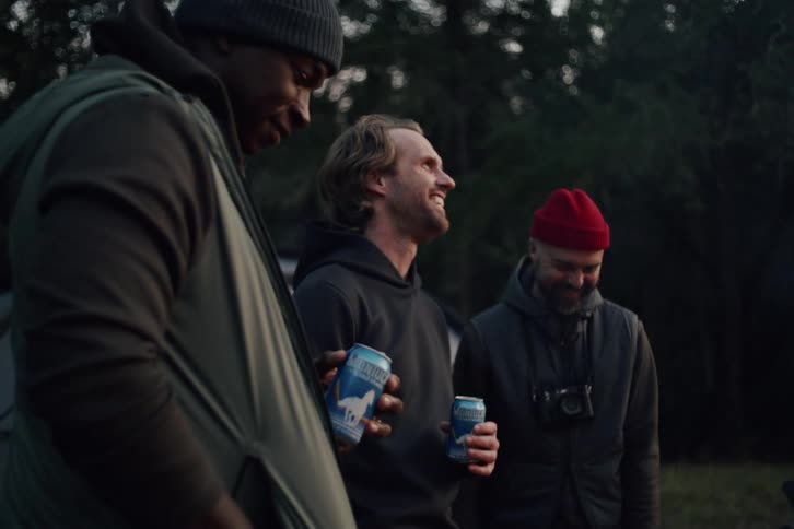 A group of diverse male friends laugh together while enjoying beers at their forest campsite at dusk