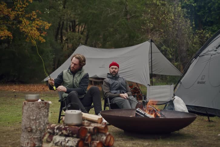 Two male friends relax at their campsite by a fire while one prepares his fly fishing rod