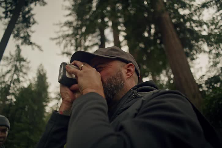 A bearded man looks through a rangefinder at the top of towering trees, then lowers it to take in the majestic view
