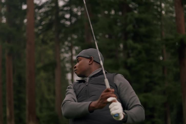 A man in warm clothing swings his golf club in slow motion, with a dense forest of tall trees in the background