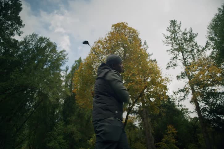 Low-angle slow motion shot of a man taking a powerful golf swing on a tree-lined course